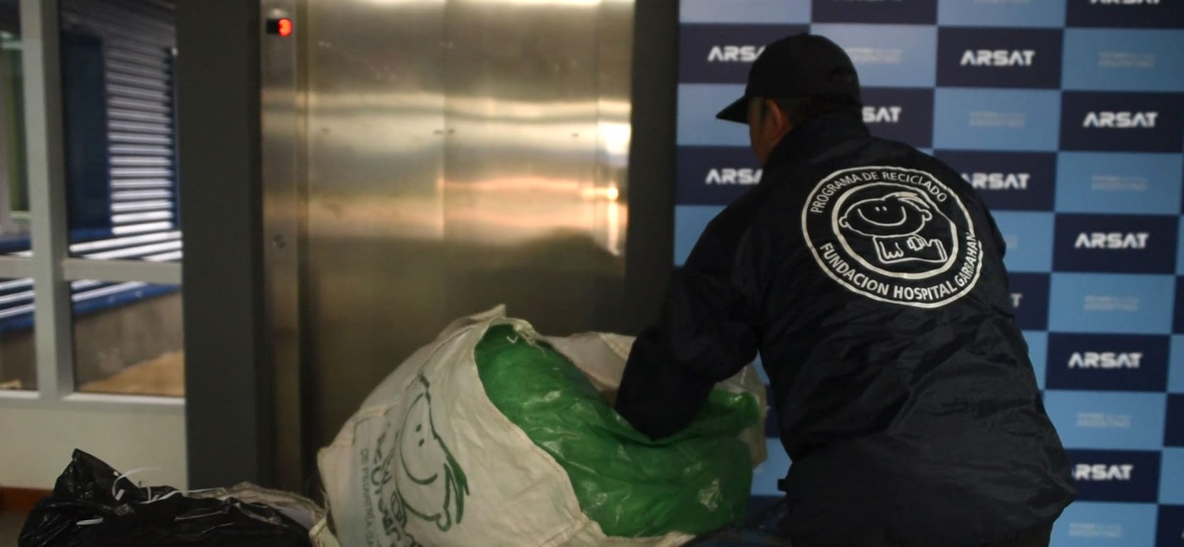 De espaldas, un hombre de la Fundación del Hospital Garraham traslada una bolsa con reciclajes, al fondo se ve un panel con logos de Arsat.