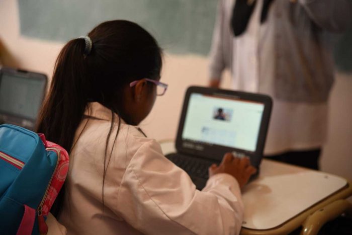 De espalda, una niña con guardapolvo y mochila utiliza una notebook en un pupitre de la escuela.