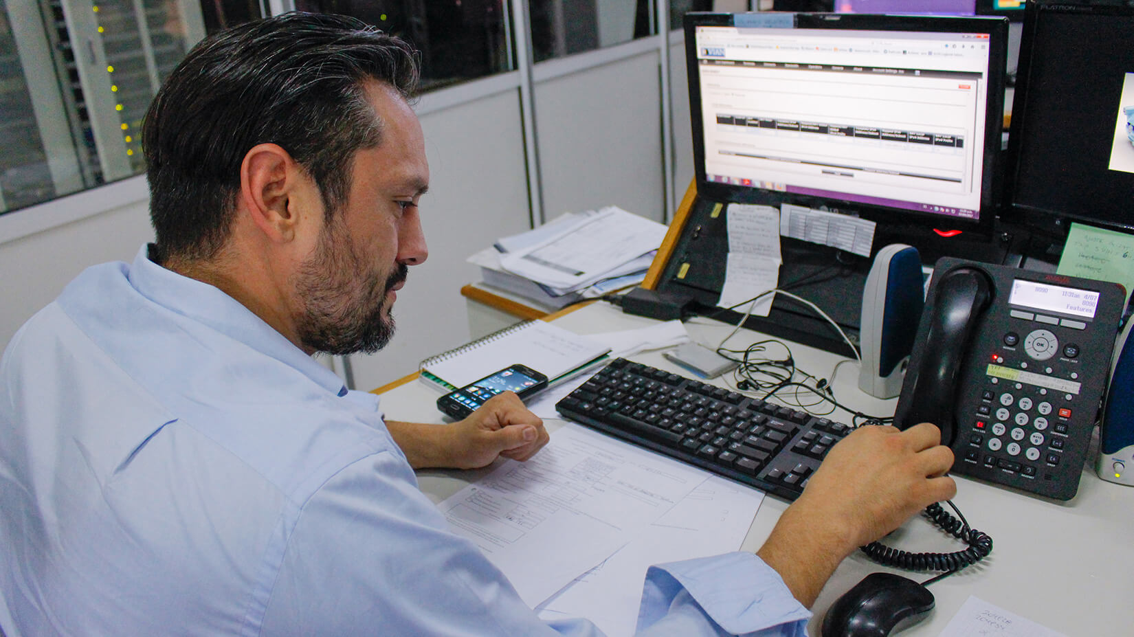 Técnico trabaja en una computadora desde el Centro de Operaciones de Benavídez. A su derecha, un teléfono fijo y a su izquierda un celular personal.