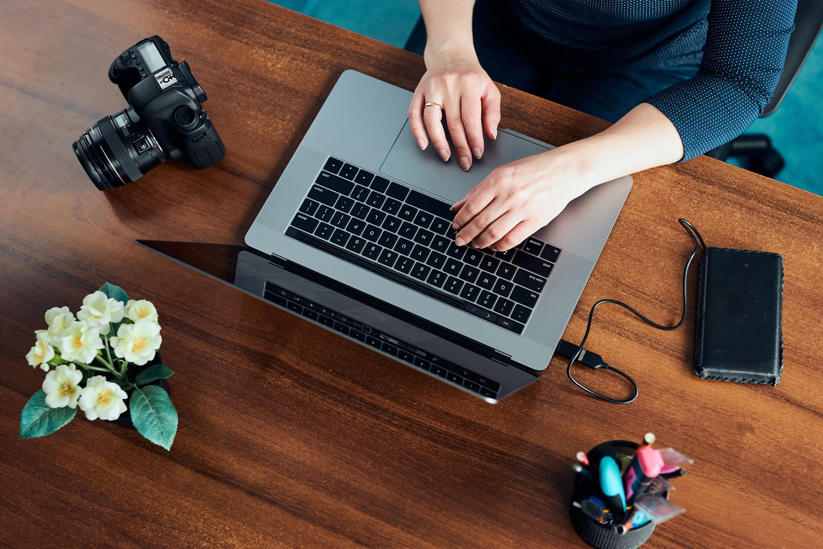 Plano cenital de manos de mujer sobre una Macbook, en su lateral izquierdo hay una cámara reflex y flores blancas y en su derecho, un disco externo.