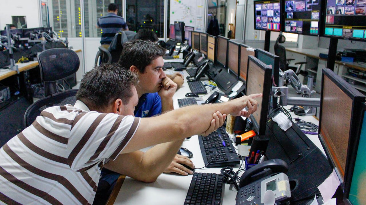Especialistas en tecnología informática, sentados con auriculares en escritorios compartidos, trabajando con una computadora y 2 pantallas