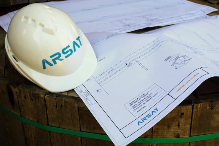 Sobre un barril de madera, hay un casco de seguridad de Arsat y un plano de arquitectura de la red federal de fibra de una cooperativa de un pueblo.