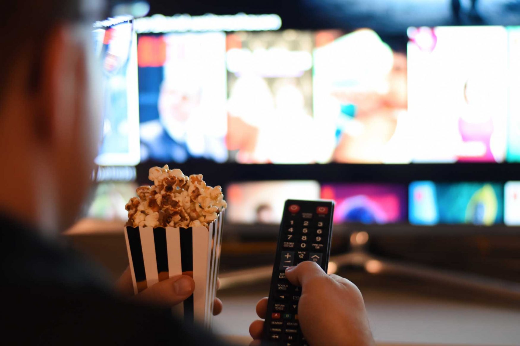 De espaldas, un hombre sostiene con una mano, una porción de pochoclos y en la otra, el control remoto que apunta a un televisor con Cinear Play.