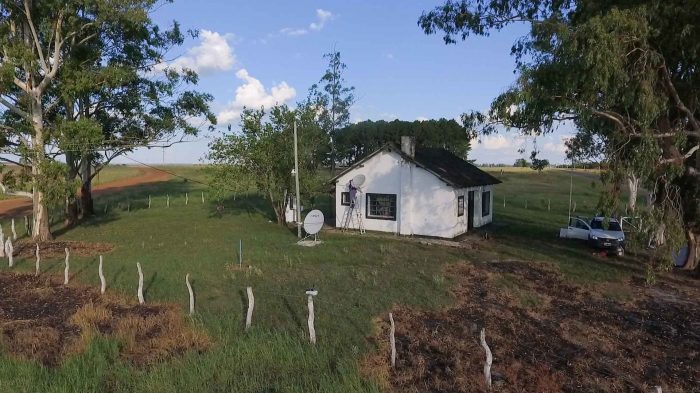 Desde un dron, una escuela rural rodeada de un campo verde. En el piso hay instalada una antena de internet y en la pared una de Televisión digital.