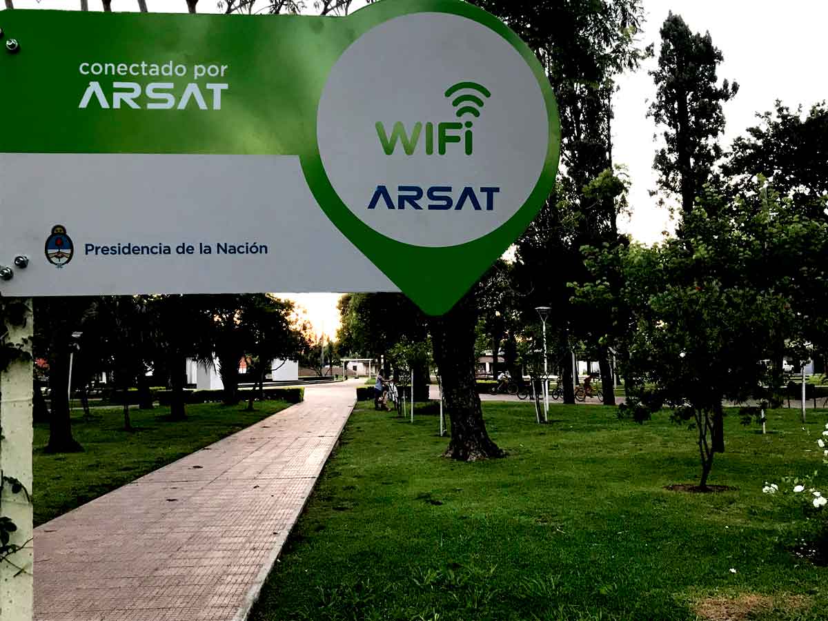 Cartel blanco y verde con logo de Arsat, de un punto wifi en la plaza de un pueblo.